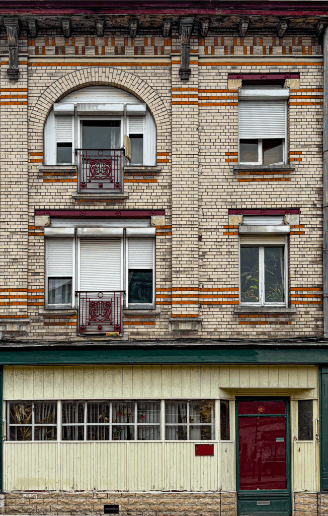 Mmodest early 20th-century residential building with a decorative brick façade in red and white. The upper floors feature arched and rectangular windows fitted with white roller shutters and framed by patterned brickwork, with wrought-iron balconies painted dark red. The cornice is supported by carved wooden brackets, adding ornamental detail. The ground floor contrasts with light vertical paneling, a row of windows with curtains, and a dark red entrance door. The overall appearance combines functional design with small decorative touches typical of working-class urban architecture.