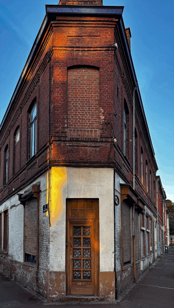 A building set at the corner of two streets, shaped like a triangle reminiscent of a “flatiron.” Its façade is made of dark brick, with several openings either bricked up or sealed with metal shutters. On the ground floor, a glass-panelled door with decorative details stands at the pointed tip of the building, framed by a lighter plaster section contrasting with the brick. Electrical cables run across the façade, and a streetlamp juts out above. On the sidewalk, trash bins and a traffic sign complete the scene. The overall impression is one of abandonment, a place where time seems to have stopped.