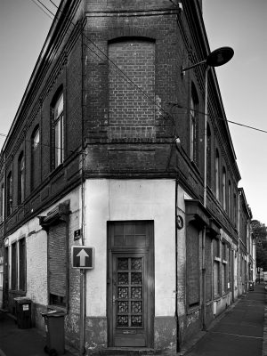 A building set at the corner of two streets, shaped like a triangle reminiscent of a “flatiron.” Its façade is made of dark brick, with several openings either bricked up or sealed with metal shutters. On the ground floor, a glass-panelled door with decorative details stands at the pointed tip of the building, framed by a lighter plaster section contrasting with the brick. Electrical cables run across the façade, and a streetlamp juts out above. On the sidewalk, trash bins and a traffic sign complete the scene. The overall impression is one of abandonment, a place where time seems to have stopped.