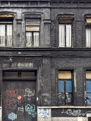 Façade d’un immeuble ancien en briques peintes en noir, à deux niveaux. En bas, une grande porte en bois et deux fenêtres, tous recouverts de graffitis et marqués par l’usure. En haut, quatre fenêtres étroites avec rideaux ou volets, encadrements dégradés et peinture écaillée.